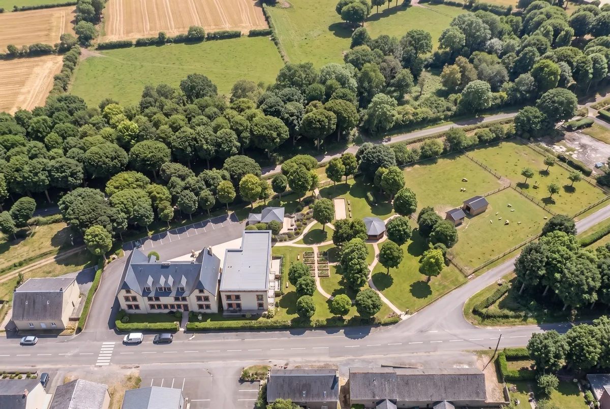 Vue aérienne de la résidence depuis la rue : le bâtiment ancien et l'aile neuve, le parc et la petite ferme derrière, la voie verte en lisière des bois.
