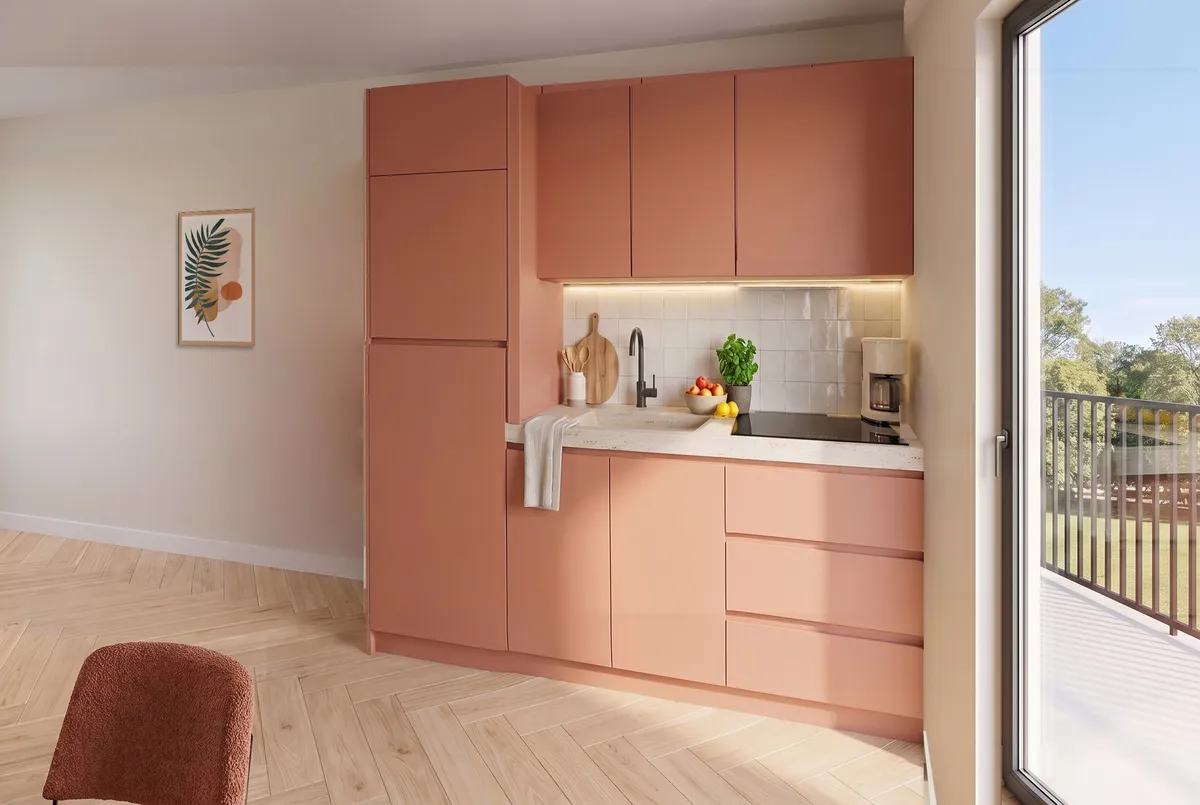 La cuisine du T2, en façades terracotta, à côté de la baie vitrée du balcon : un évier, une plaque de cuisson, des tiroirs, des meubles hauts et une grande colonne.